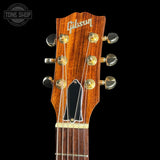 Close-up of a Gibson acoustic guitar headstock with six gold tuning pegs, a black pickguard, and a warm brown wood finish, shown against a black background with a Tone Shop watermark in the top left.
