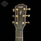 Close-up of a Taylor acoustic guitar headstock with six gold tuning pegs, six strings, and a dark brown wood finish against a black background.