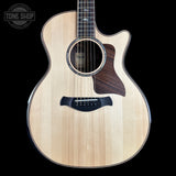 A Taylor acoustic-electric guitar with a natural wood finish and dark brown pickguard, shown against a solid black background with a Tone Shop watermark in the top left corner.