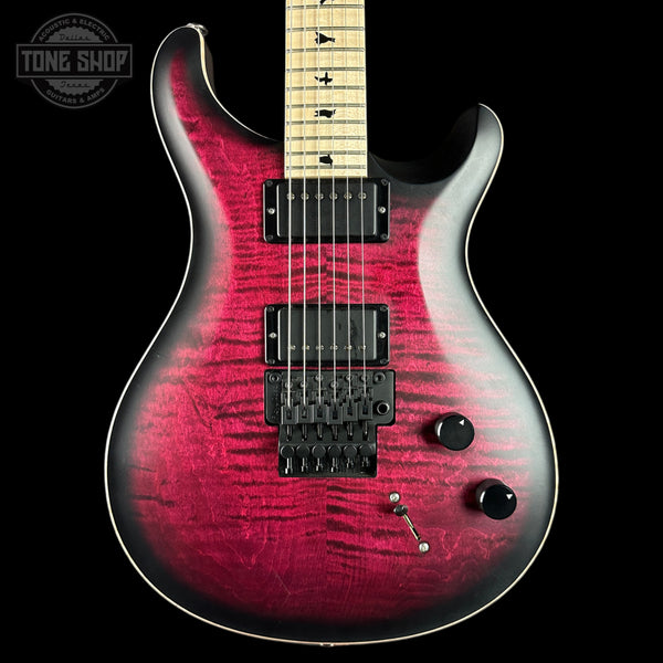 A double-cutaway electric guitar with a vibrant magenta-pink to black flame finish on a black background. The neck is light wood with black inlays.