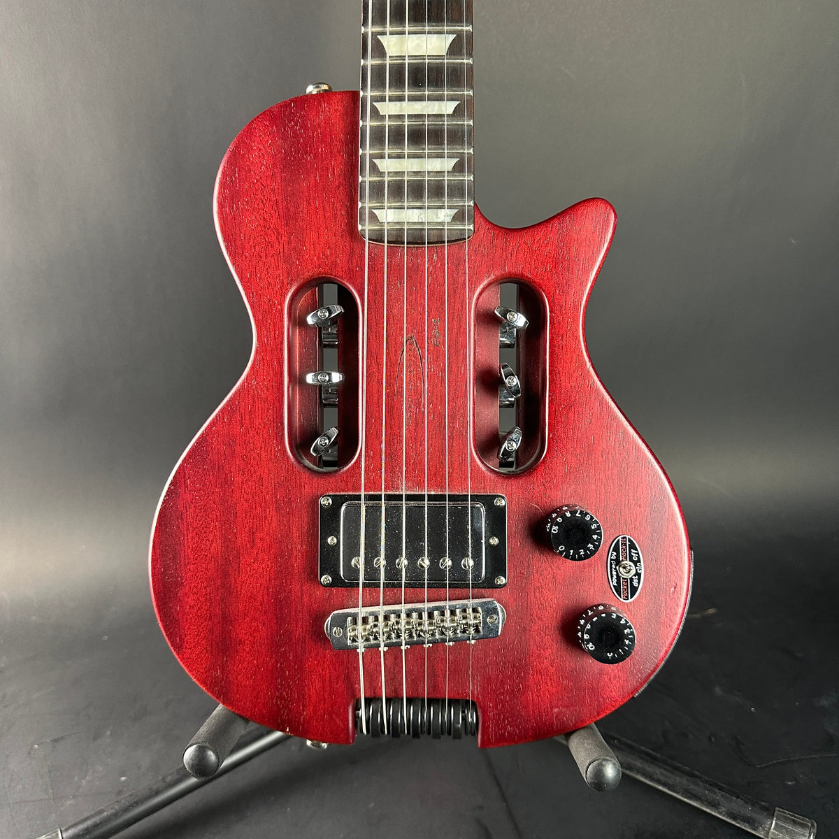 A red electric guitar with a double-cutaway body, dual humbucker pickups, and black control knobs, displayed on a stand against a dark gray background.