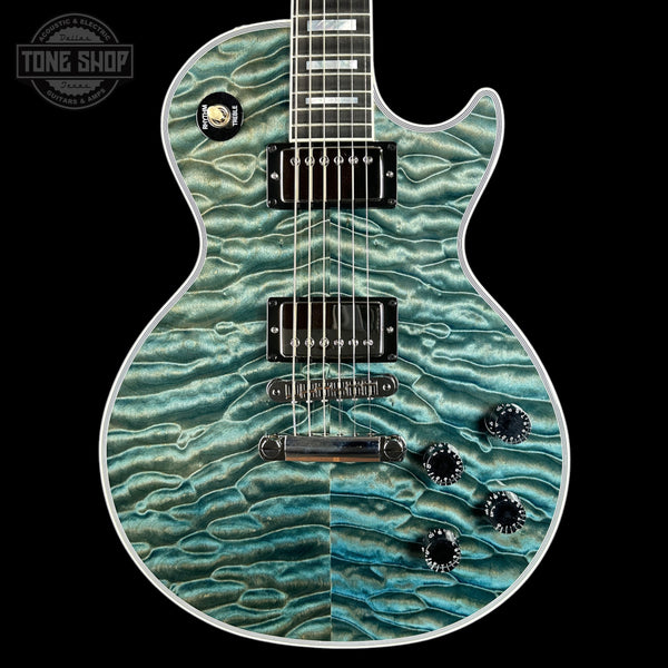 A double-cutaway electric guitar with a vibrant teal and turquoise flamed maple top, featuring black hardware and control knobs, set against a solid black background with a Tone Shop watermark in the upper left corner.