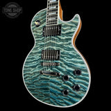 A double-cutaway electric guitar with a vibrant turquoise and teal flamed maple top, black hardware, and a natural wood neck and body edge, shown against a solid black background with a Tone Shop watermark in the top-left corner.