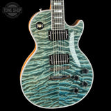 A close-up of an electric guitar body featuring a vibrant turquoise and teal wavy quilted flamed maple top, with a black pickguard, chrome hardware, and a dark fretboard. The Tone Shop logo is visible in the top-left corner.