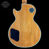 The back of an electric guitar with a natural wood finish, featuring a black pickguard and a blue circular 'Gibson' logo badge on the upper right side, set against a solid black background.