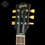 Close-up of the headstock of a Gibson Les Paul Custom electric guitar, featuring a black headstock with gold Gibson and Les Paul Custom lettering, six yellow tuning pegs, and a dark wood fretboard, set against a solid black background.
