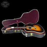 An acoustic guitar with a sunburst finish displayed inside an open hard case with a burgundy velvet interior, set against a solid black background.