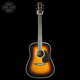 A classic acoustic guitar with a sunburst finish, shown vertically against a solid black background. The guitar features a dark brown outer edge fading to a warm golden-orange center, with a black pickguard and white binding.
