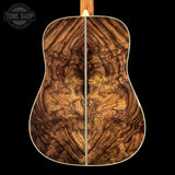 The back of an acoustic guitar featuring an intricate, highly figured wood top with swirling grain patterns in shades of brown and golden tan, set against a solid black background.