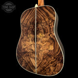 Close-up of the back of an acoustic guitar showing a highly figured brown wood top with dramatic swirling grain patterns, white binding, and a black side body, set against a solid black background with a Tone Shop watermark in the top left.