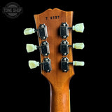 Close-up of the headstock of a guitar, showing six tuning pegs, black tuning machines, and the serial number 76727 on a warm brown wood finish, set against a solid black background.