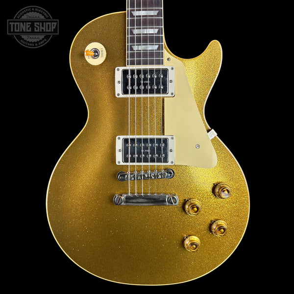 A gold glitter electric guitar with a cream pickguard, chrome hardware, and a dark fretboard, shown against a solid black background. A Tone Shop watermark is visible in the top-left corner.