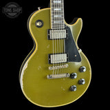A Gibson Les Paul Custom electric guitar with a distressed olive green finish, black pickguard, and chrome hardware, shown against a solid black background with a Tone Shop watermark in the top left corner.