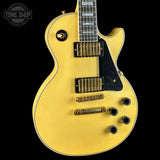 A yellow-finish Gibson Les Paul Custom electric guitar with a black pickguard, gold hardware, and black control knobs, shown against a solid black background with a Tone Shop watermark in the top left corner.