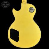 The back of a Gibson electric guitar in a vibrant matte yellow finish, shown against a solid black background. The Gibson logo is visible on the upper back, and a black pickguard is located on the lower left.