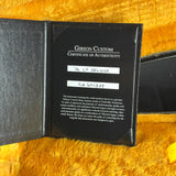 An open black Gibson Custom Certificate of Authenticity booklet resting on a bright yellow faux fur surface. The certificate is addressed to '76 LP DELUXE and includes handwritten labels.