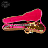 A gold-finish electric guitar resting inside an open hard case with bright pink velvet lining, shown against a black background. A Tone Shop watermark is visible in the upper left corner.