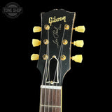 Close-up of a Gibson Les Paul Custom electric guitar headstock, featuring a black wood finish with gold hardware and yellow tuning pegs, set against a solid black background.