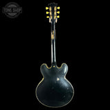 A vintage black electric guitar shown from the back against a solid black background, featuring gold tuning pegs and gold edge trim, with a Tone Shop watermark in the top left corner.