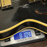 Close-up of a black electric guitar body with a gold outline, resting on a digital kitchen scale displaying 8 lbs 10.4 oz.