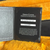 An open Gibson Custom Certificate of Authenticity booklet placed on a yellow faux-fur surface. The black certificate insert shows the text 'Gibson Custom Certificate of Authenticity' and handwritten instrument details, with part of a black case visible on the right.