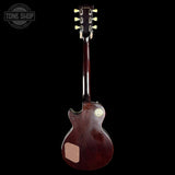 A dark brown electric guitar shown from the back against a solid black background, featuring a glossy finish, a light wood pickguard, and a gold-toned circular badge on the body.
