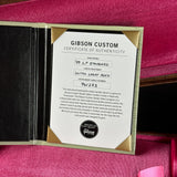 A Gibson Custom Certificate of Authenticity card displayed inside an open presentation case on a pink textured surface. The card is white with black text and gold border accents, and the case interior is black.