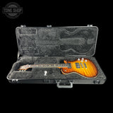 An electric guitar with a sunburst finish resting inside an open black hard case with a gray padded interior, shown against a solid black background. A Tone Shop logo watermark is visible in the top-left corner.