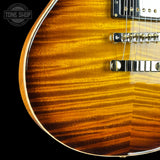 Close-up of a double-cutaway electric guitar body showing a flamed maple wood finish in a warm amber-brown sunburst, with chrome hardware and a black pickguard against a black background. A Tone Shop watermark is visible in the top-left corner.
