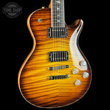 A close-up of an electric guitar body showing a vibrant sunburst finish that transitions from bright amber-orange to dark brown wood grain. The glossy finish reflects light, and chrome hardware is visible against the black background.