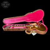 A gold electric guitar inside an open hard case with a bright pink velvet interior, shown against a black background. A Tone Shop watermark is visible in the top-left corner.