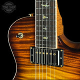 Close-up of an electric guitar body and neck showing a flamed maple wood finish in a warm brown sunburst, with a black humbucker pickup and bird inlays on the fretboard.