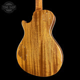 The back of an electric guitar with a rich, warm honey-amber wood finish and prominent grain, shown against a solid black background. A Tone Shop watermark is visible in the top-left corner.