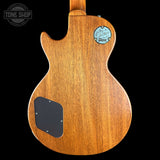 The back of a Gibson electric guitar with a natural wood finish, featuring a black pickguard and a circular blue Gibson logo badge on a black background.