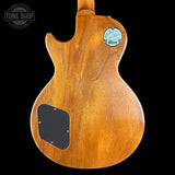 The back of a Gibson Les Paul Custom electric guitar, featuring a natural golden-brown wood finish, a black pickguard, and a circular blue-and-white Gibson logo badge on the upper back, set against a solid black background.