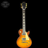 A Gibson Les Paul Custom electric guitar with a natural flame maple top, shown against a solid black background. The guitar features a black headstock with gold Gibson branding, a dark fretboard with gold block inlays, and gold hardware.