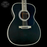 A close-up studio shot of the lower half of an acoustic guitar against a black background. The guitar has a glossy black body, a dark pickguard, and a silver-toned bridge with gold-colored tuning pegs.