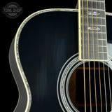 Close-up of a black acoustic guitar body with iridescent inlay around the soundhole and edge, a light wood fretboard, and colorful mother-of-pearl style fretboard inlays. A Tone Shop watermark is visible in the top left.