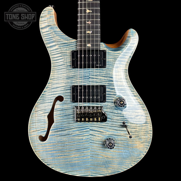 A close-up of an electric guitar body with a light blue and white flamed maple wood finish, featuring black hardware and pickups, set against a solid black background with a Tone Shop watermark in the top-left corner.