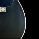 Close-up detail of a black electric guitar body showing the edge of the soundhole surround, a silver metal bridge plate with a gold-colored inlay, and a black background.