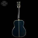 The back of an acoustic guitar with a glossy black finish and a single light-colored wood spine, shown against a solid black background. A Tone Shop watermark is visible in the top-left corner.