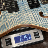 A close-up of a blue flamed maple electric guitar body resting on a digital scale displaying 6 lbs 15.7 oz.