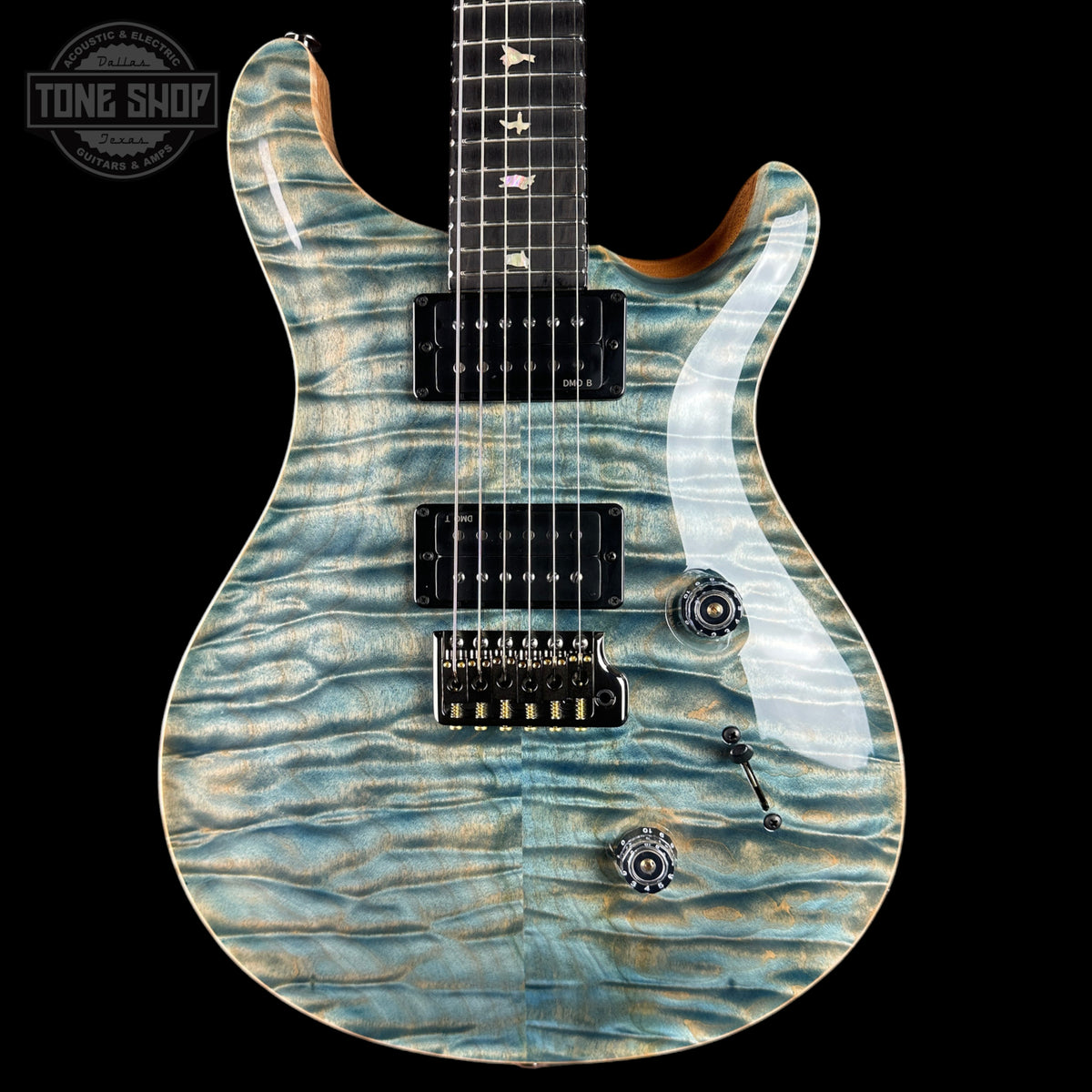 A close-up of an electric guitar body with a blue and gray wavy maple wood finish, featuring black hardware and a black pickguard, shown against a solid black background with a Tone Shop watermark in the top left corner.