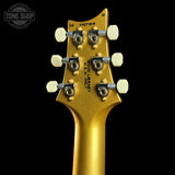 Close-up rear view of a gold electric guitar headstock with six white tuning pegs and black adjustment screws, shown against a black background. A Tone Shop watermark is visible in the upper left corner.
