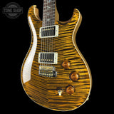 A close-up of a PRS electric guitar body featuring a vibrant flame maple finish with a golden-brown and amber zebra wood grain pattern, set against a solid black background.