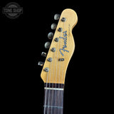 Close-up of the headstock of a Fender Telecaster electric guitar against a solid black background, showing the natural wood headstock, six silver tuning pegs, and the blue Fender logo.