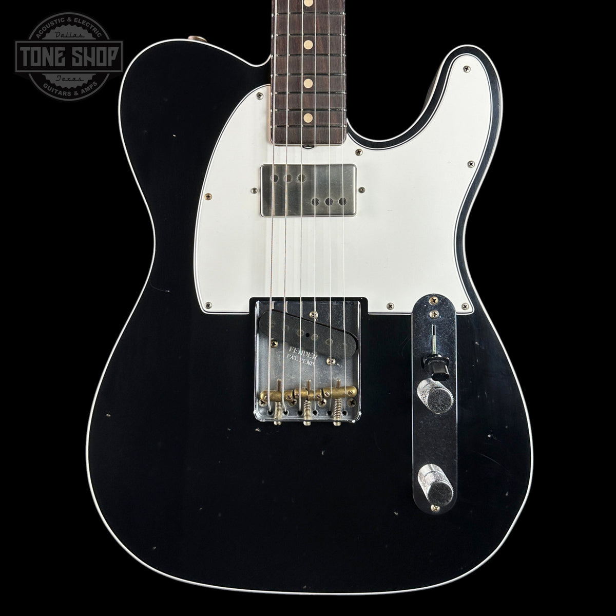 A black electric guitar with a white pickguard, featuring a dark fretboard and gold-toned hardware, shown against a solid black background with a Tone Shop watermark in the top-left corner.