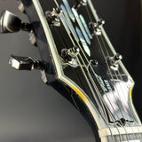 Close-up of the headstock of a black electric guitar, showing the tuning pegs, strings, and white 'Colomb' branding on a dark gray background.