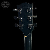 Close-up of the headstock of a black electric guitar against a black background, showing six black tuners with silver screws and a visible crack near the lower edge. A Tone Shop watermark is in the top left corner.