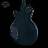 The back of a black electric guitar body, photographed against a solid black background. The glossy finish shows some light dust and scratches, and a circular Tone Shop watermark is visible in the top-left corner.
